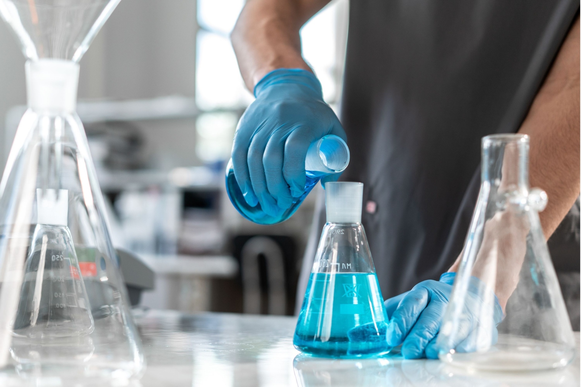 Chemikalienschutzhandschuhe, die im Bereich Chemie genutzt werden und eine Flüssigkeit von einen Erlenmeyerkolben in einen anderen kippt