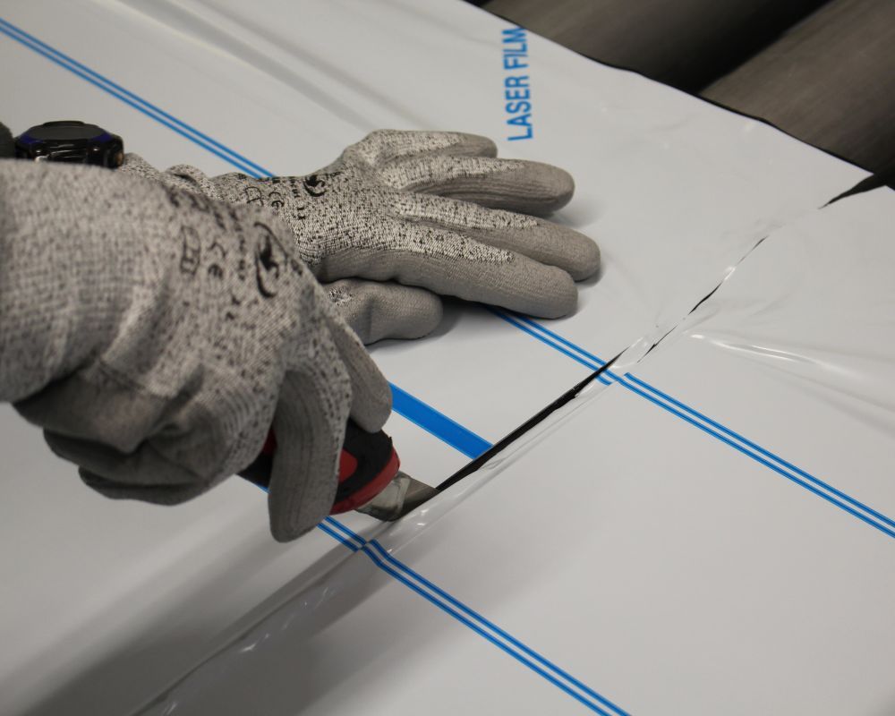 Arbeiter schneidet mit einem Cuttermesser in eine Folie. Dabei trägt er Schnittschutzhandschuhe für seinen Schutz